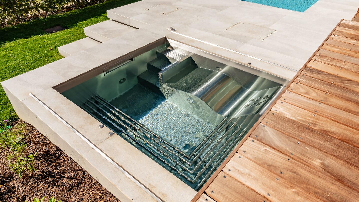 Stainless steel jet spa pool set into a stone and cedar deck in a private backyard, with welded bench seating and a water feature